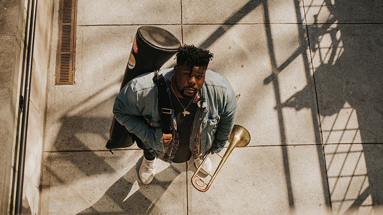 Jeffrey Miller looks up at the camera from a sidewalk holding his trombone